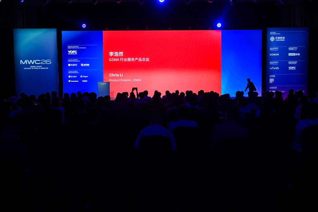 A dark auditorium is filled with seated attendees facing a brightly lit stage. The screen displays a red and blue background with the name “Chris Li, Product Director, GSMA.” Side banners show "MWC26" and sponsor logos. Silhouettes of people are visible.
