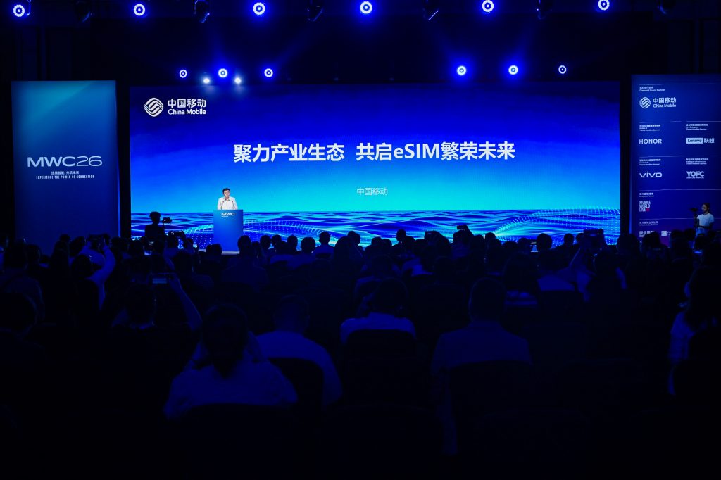 A speaker stands at a lectern on stage at a tech conference, with a large blue screen displaying Chinese text and “eSIM.” The audience sits in a dimly lit room. The event is hosted by China Mobile, and banners for MWC26 and tech brands are visible.
