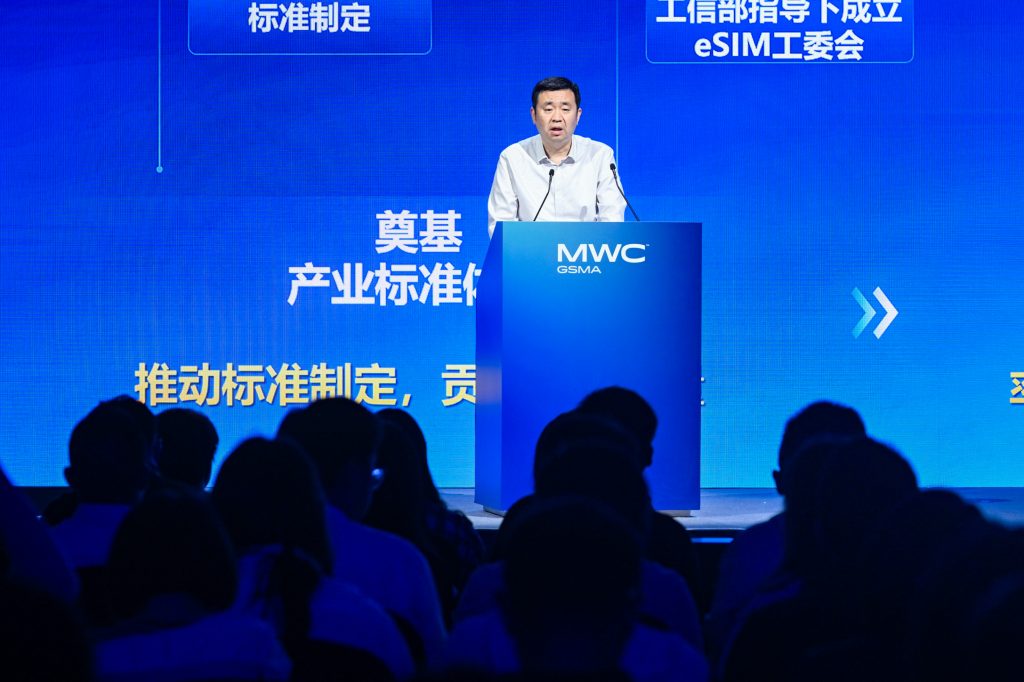 A man in a white shirt stands at a blue podium labelled "MWC GSMA," speaking to an audience. Blue screens with white Chinese and English text form the background, highlighting topics like eSIM and industry standards. Audience members are mostly silhouettes.