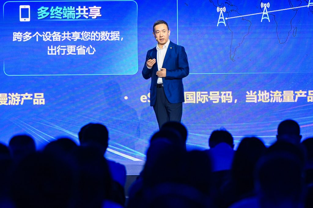 A man in a blue suit speaks on stage, holding a remote. Behind him is a blue screen with Chinese text and technology graphics. An audience faces him, silhouetted in the foreground, listening to the presentation.