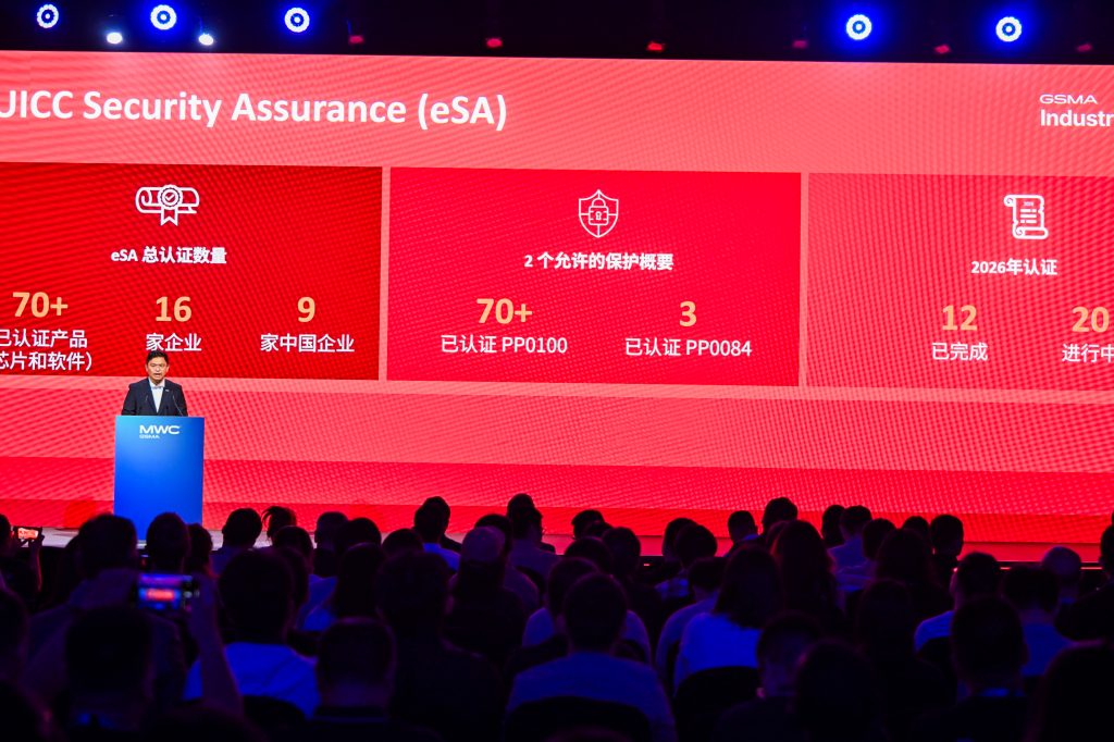A speaker stands at a lectern onstage before an audience. Behind him is a large red screen displaying statistics about "eSA Security Assurance" with white icons, text, and numbers, indicating certifications, companies involved, completed projects, and 2026 targets.