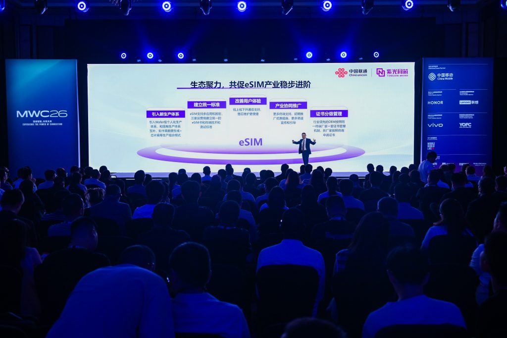 Audience facing a large screen at a technology conference, MWC26. The screen displays a presentation about eSIM in Chinese with charts and company logos. Blue stage lights illuminate the room, and attendees are seated in rows, watching the presentation.