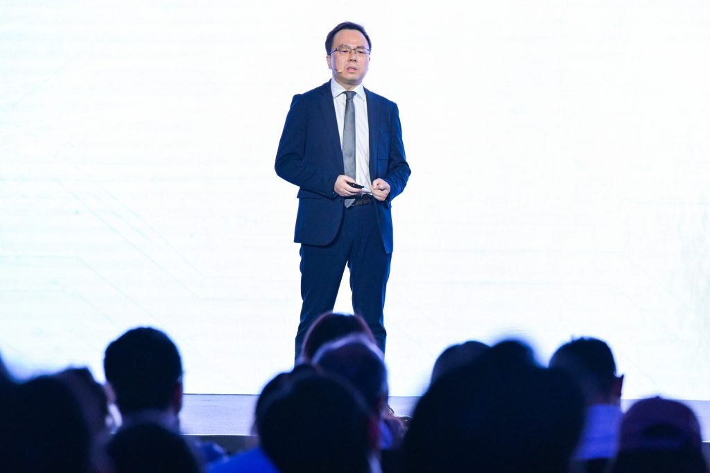 A man in a dark suit, white shirt, and tie stands on stage in front of a bright, white background, holding a small object. He faces an audience, seen in silhouette in the foreground. The setting appears to be a formal presentation or conference.