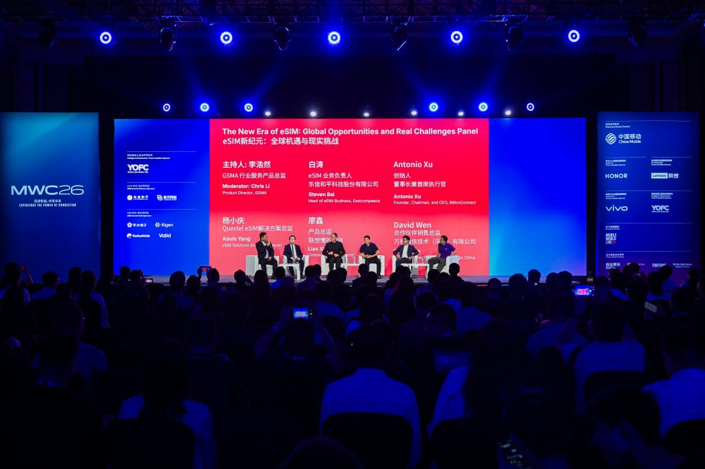 A large audience watches a panel of five people seated on stage at MWC26, with a red background screen displaying panellists' names and discussion topic in English and Chinese. Blue lighting illuminates the venue, and many attendees are taking photos or videos.