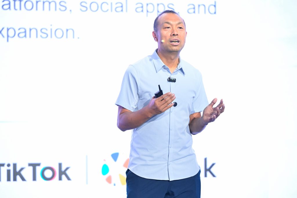 A man with short hair, wearing a light blue short-sleeved shirt, speaks on stage holding a marker. He has a headset microphone. Behind him, a large screen displays partial text and TikTok logos in a bright, well-lit setting.