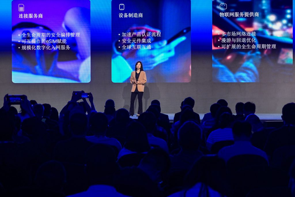 A woman stands on stage giving a presentation to an audience. Behind her are three large screens with text and images in Chinese, highlighting technology services. Audience members are seated, some holding up phones to take photos or record the event.
