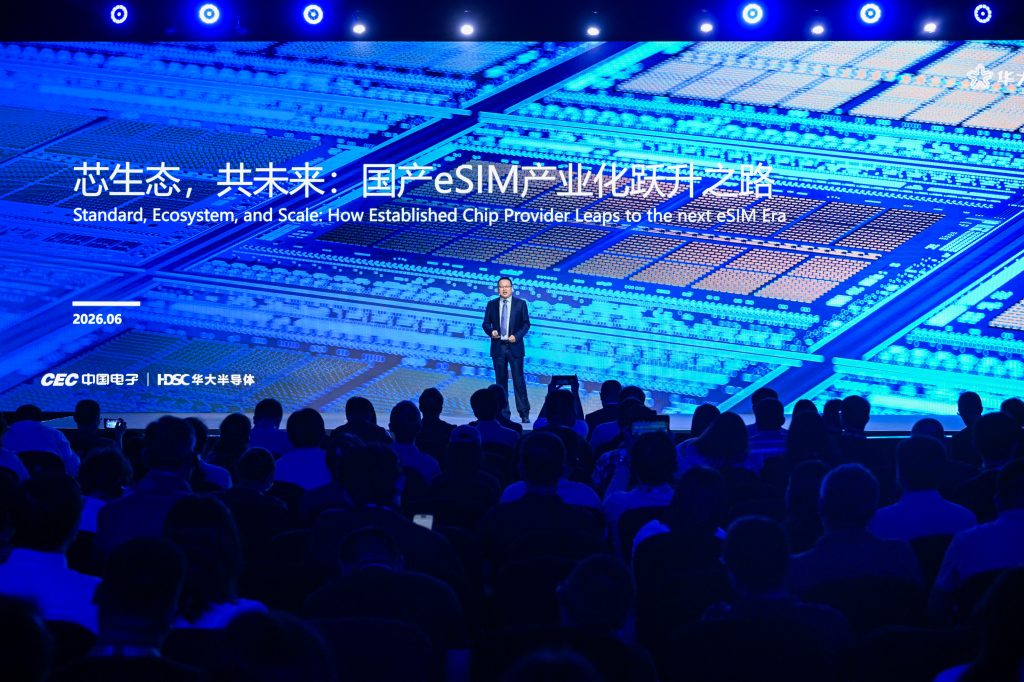 A man in a suit stands on stage in front of a large blue microchip graphic and bilingual text about eSIM technology. An audience watches, seated in a dark auditorium. The event date is June 2026, and the screen shows English and Chinese text.