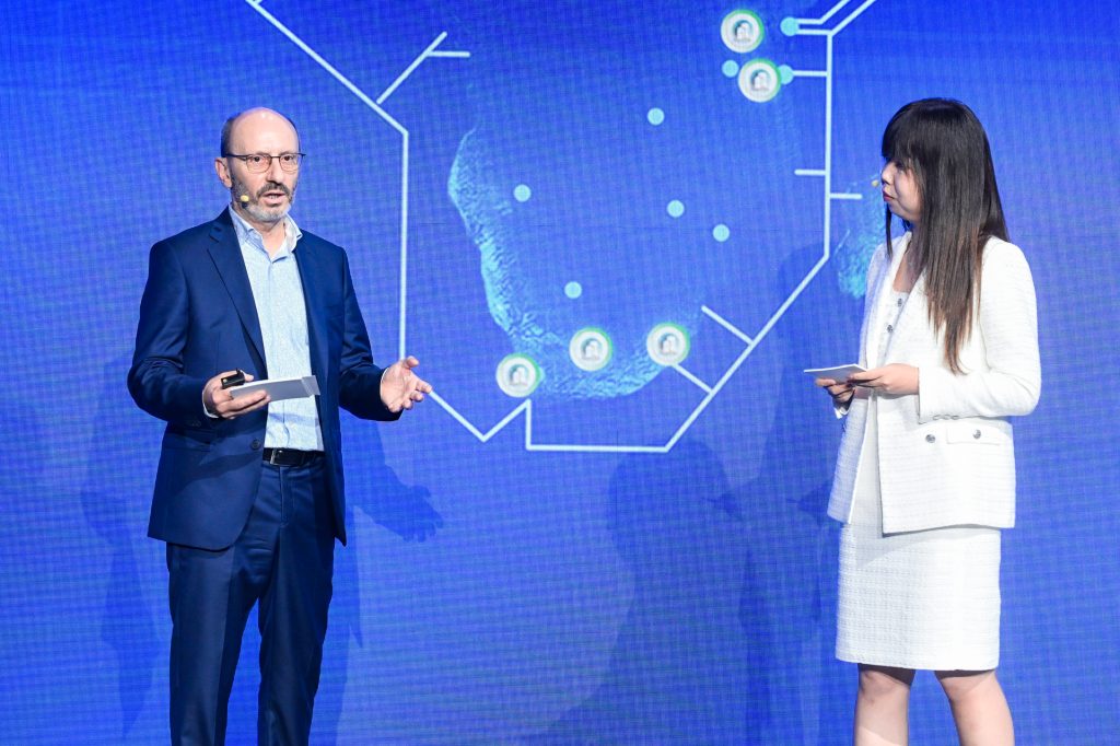 A man in a blue suit speaks whilst holding a mobile on stage next to a woman in a white suit holding a tablet. They stand before a blue screen displaying a map with interconnected icons. Both appear engaged in conversation.
