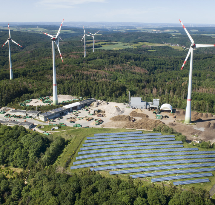 Aerial view of a renewable energy site featuring four wind turbines and an extensive solar panel array. The area is surrounded by dense green forests and rolling hills under a clear sky. Nearby, industrial buildings and construction materials are visible amidst cleared land.