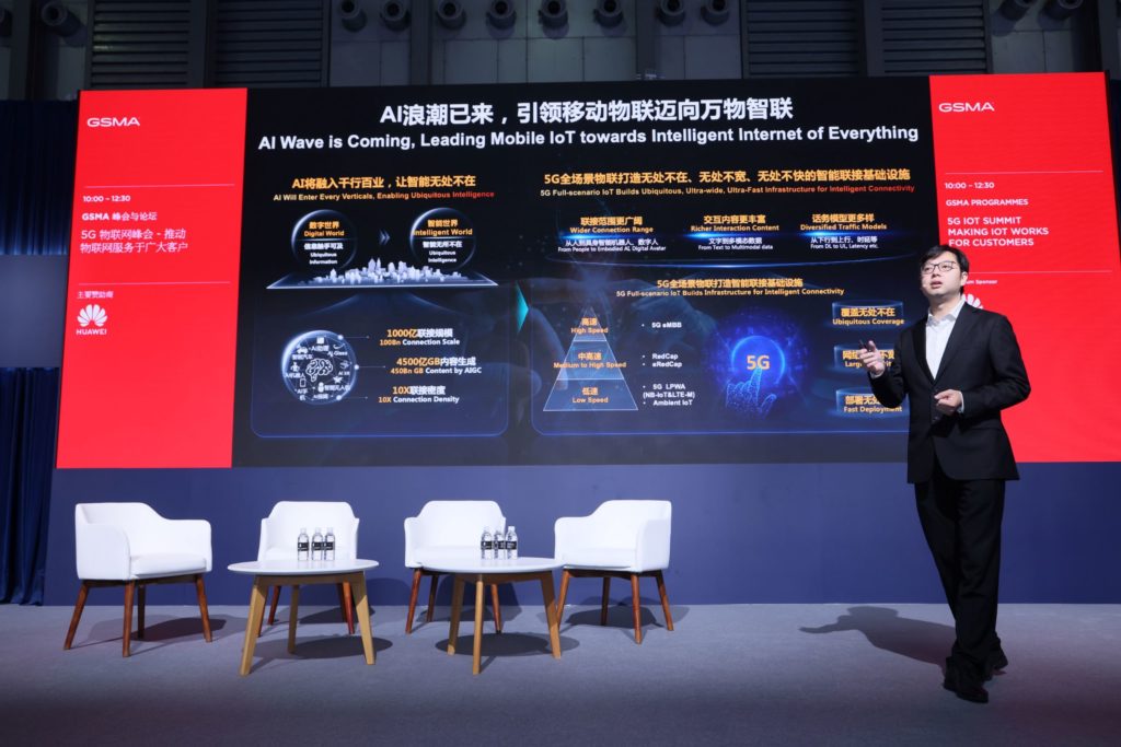 A man speaking on stage with a big screen behind him with text and images and 4 chairs and 2 tables on the stage.