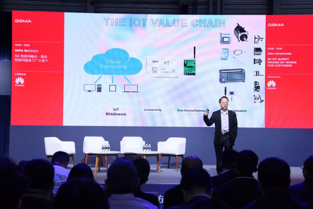 A man standing on stage talking with a big screen behind him with images