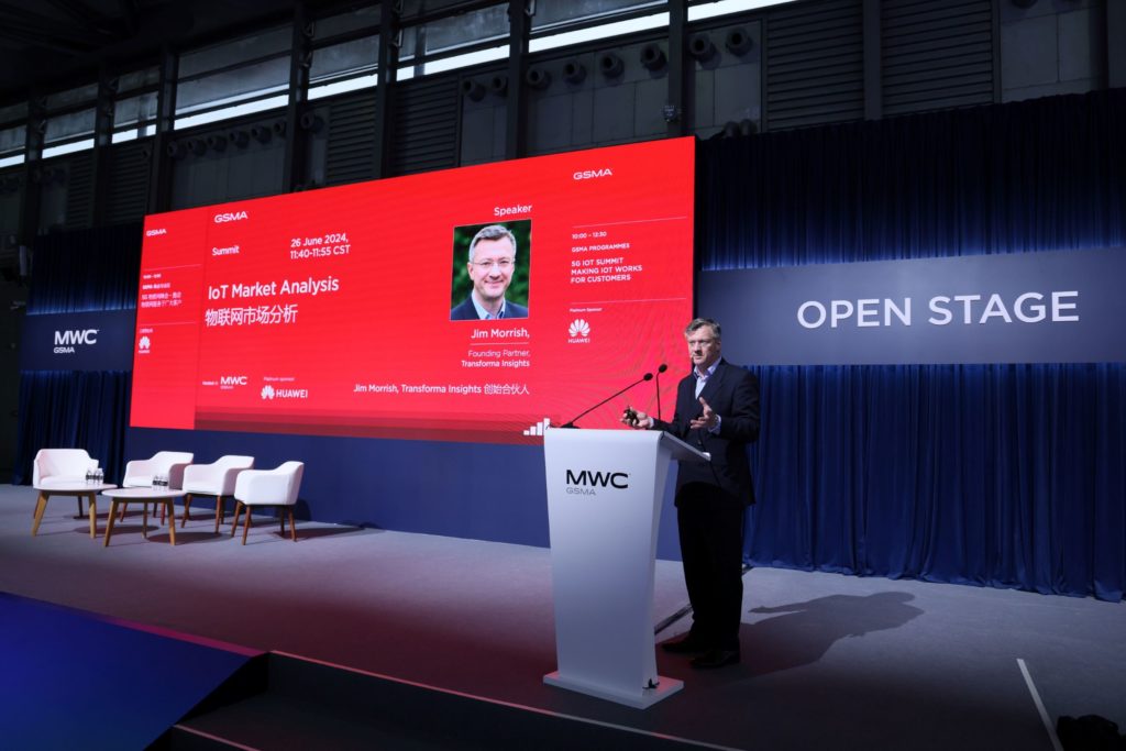 A man speaking on stage in front of a podium with a big screen behind him