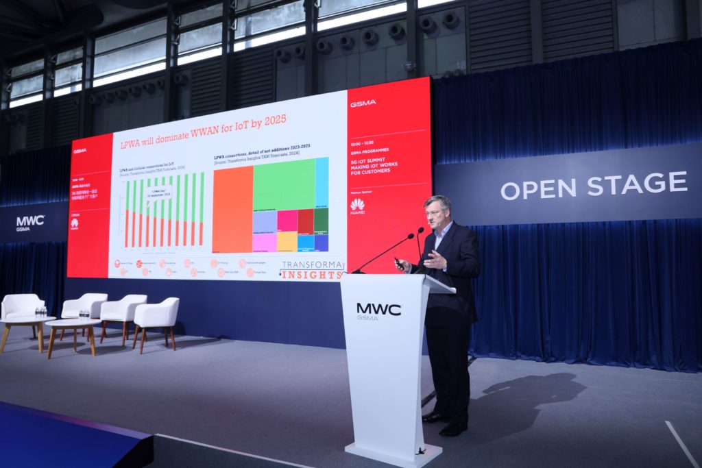 A man speaking on stage in front of a podium with a big screen behind him