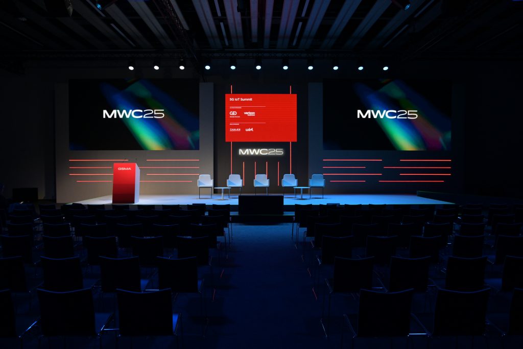 A modern conference stage with five white chairs, a red podium on the left, and three screens displaying "MWC25" with colorful graphics. The audience area has many empty black chairs, and blue lighting illuminates the space.