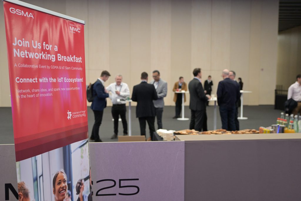 A networking event with small groups of people conversing near high tables. In the foreground, a red banner invites attendees to a networking breakfast about IoT. Food and drinks are set on tables, and the event space has plain walls and minimal decor.