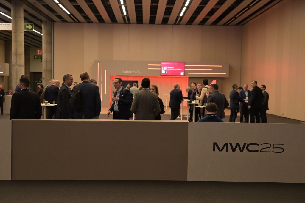 A group of people in business attire stand and converse in a modern conference hall with high ceilings. In the foreground, a partition with “MWC25” printed on it is visible. A digital screen and event branding appear in the background.