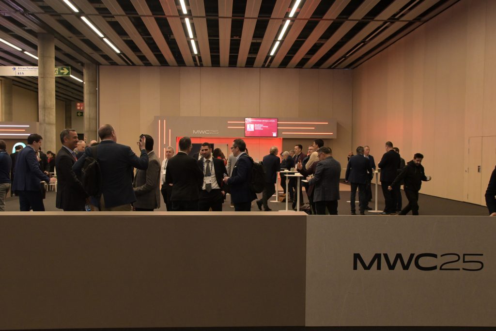 A group of people in business attire converse and network at an indoor event with minimalist decor. A large sign in the foreground reads “MWC25.” In the background, a digital screen displays event information against beige panel walls.