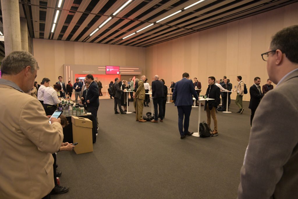 Business professionals in suits network and chat in a spacious, modern conference hall with beige walls, cocktail tables, and a refreshments area. Some people stand in groups, while others use their phones. A red-lit display is visible at the back.