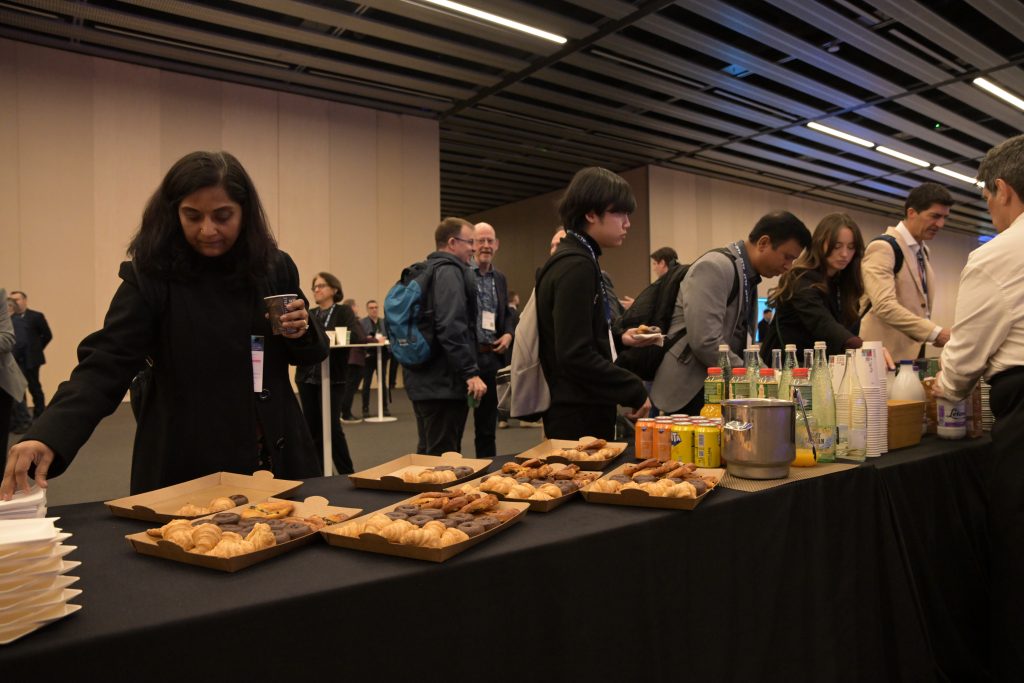 People at a conference gather around a refreshment table with pastries, bottled drinks, and cups. A woman in the foreground picks up a plate and cup, while others select snacks. The setting is a modern, spacious room with overhead lights.