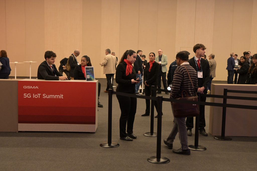 People register at a red and white desk labeled "5G IoT Summit" in a conference hall. Staff in black suits with red scarves assist attendees. A few people wait in line, while others converse in the background near beige walls.