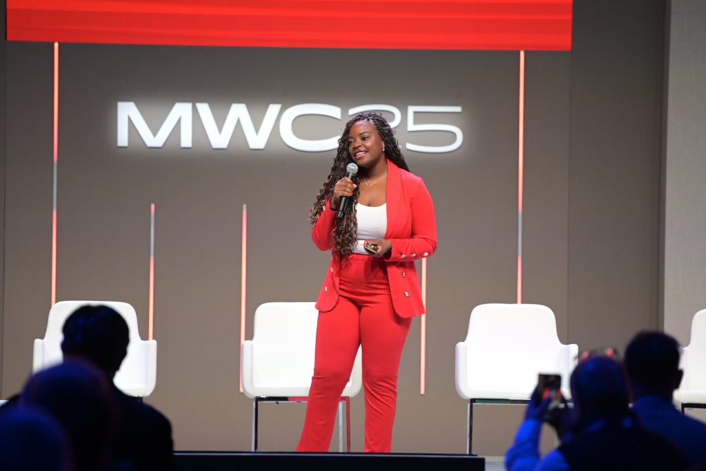 A woman in a red suit stands on stage, holding a microphone and smiling while speaking. She stands in front of a large screen displaying “MWC25.” Three empty white chairs are behind her, and audience members are visible in the foreground.