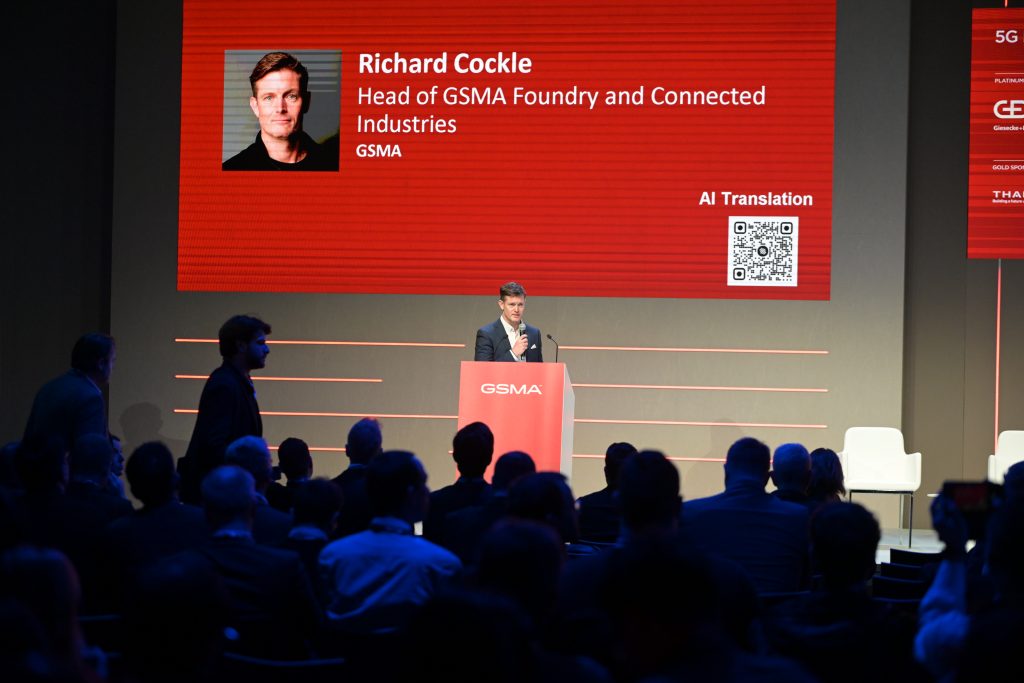 A man stands at a red podium labeled "GSMA," giving a presentation to a seated audience. Behind him is a large screen displaying his name, photo, and title: "Richard Cockle, Head of GSMA Foundry and Connected Industries," with an "AI Translation" QR code.