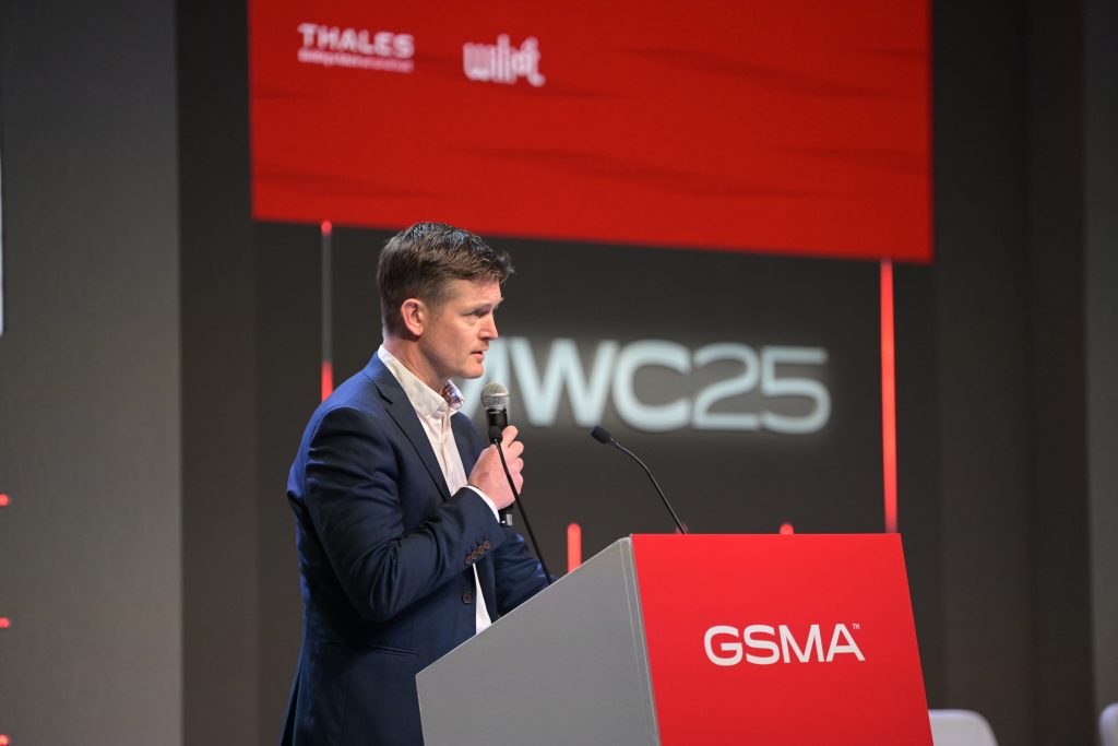 A man in a dark suit speaks into a microphone at a podium labeled “GSMA.” Behind him, a red screen displays the logos for Thales and Wiliot, and the text “MWC25” in white. The setting appears to be a professional conference or presentation.