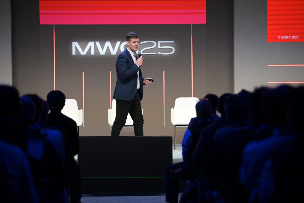 A man in a suit stands on stage holding a microphone, speaking to an audience at MWC25. Two empty white chairs are behind him. The event logo "MWC25" is illuminated on a dark backdrop with red and white lights overhead.