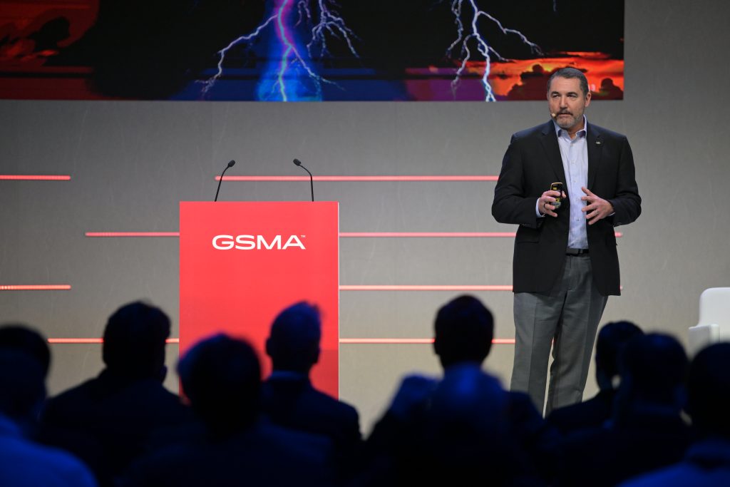 A man in a suit stands on stage holding a clicker, speaking to an audience. A red podium labeled “GSMA” is to his left. Behind him is a large screen displaying an image with blue lightning. The audience is seated in the foreground, facing the stage.
