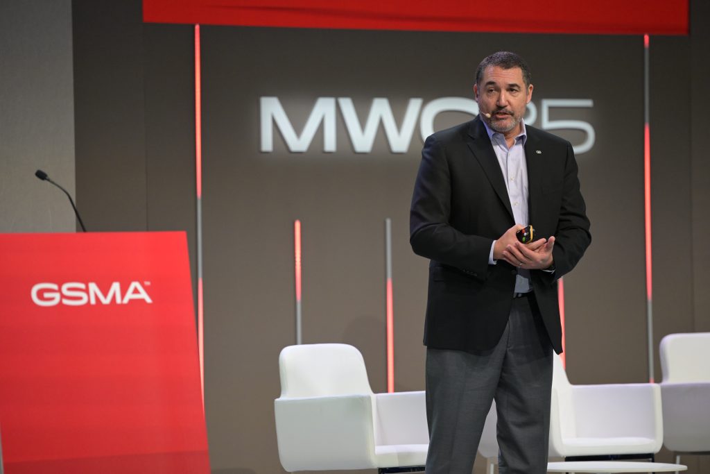 A man in a dark suit stands on stage holding a small object, speaking at the MWC23 event. Behind him are white chairs and a backdrop displaying "MWC23". To his left, there is a red podium with "GSMA" written on it.