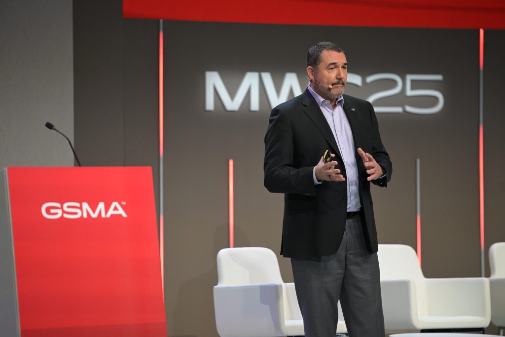 A man in a dark suit speaks on stage at MW25, standing near a podium with a red GSMA sign. He gestures with his hands and white chairs are visible behind him against a brown and red-lit backdrop.