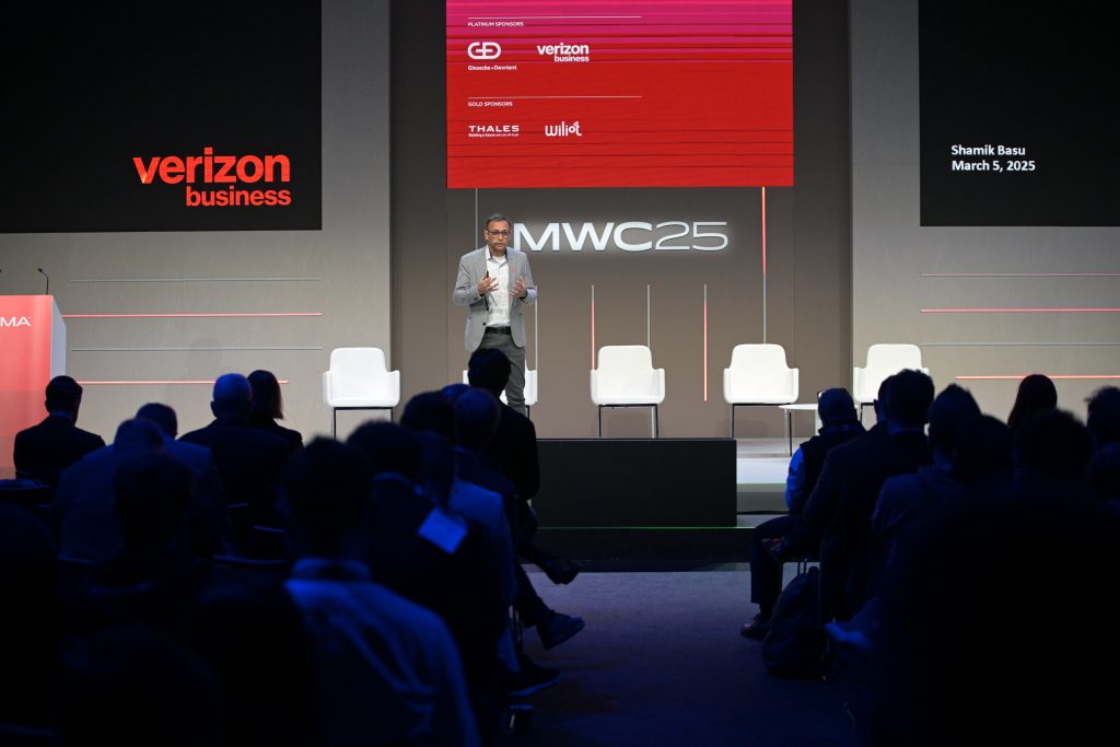 A man in a gray suit stands on stage speaking at a tech conference. The stage has four white chairs and large signs read “verizon business” and “MWC25.” Audience members sit in rows, facing the speaker, under dim lighting.