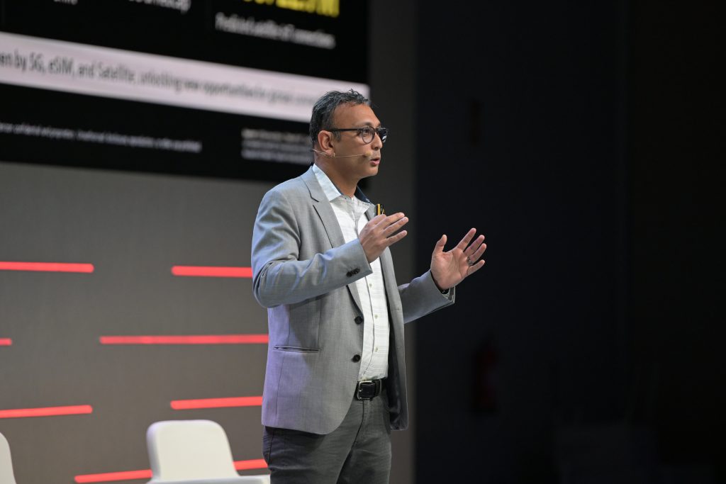 A man in a gray suit jacket, white shirt, and glasses stands speaking on stage. He gestures with open hands, appearing engaged. Behind him is a large screen with text, and to his side is a white chair. The setting suggests a formal presentation or conference.