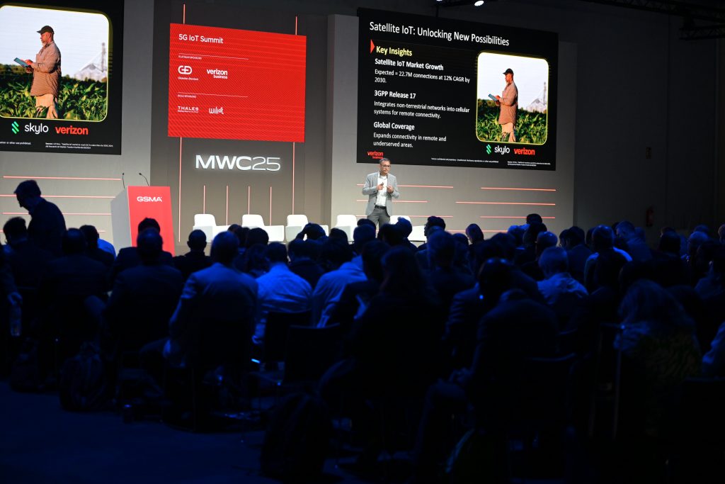 A presenter stands onstage at the MWC25 event, addressing a seated audience. Behind him is a large screen displaying a slide on “Satellite IoT,” featuring bullet points and logos for GSMA, Verizon, Skylo, and UKT, with a photo of a person holding a tablet in a field.