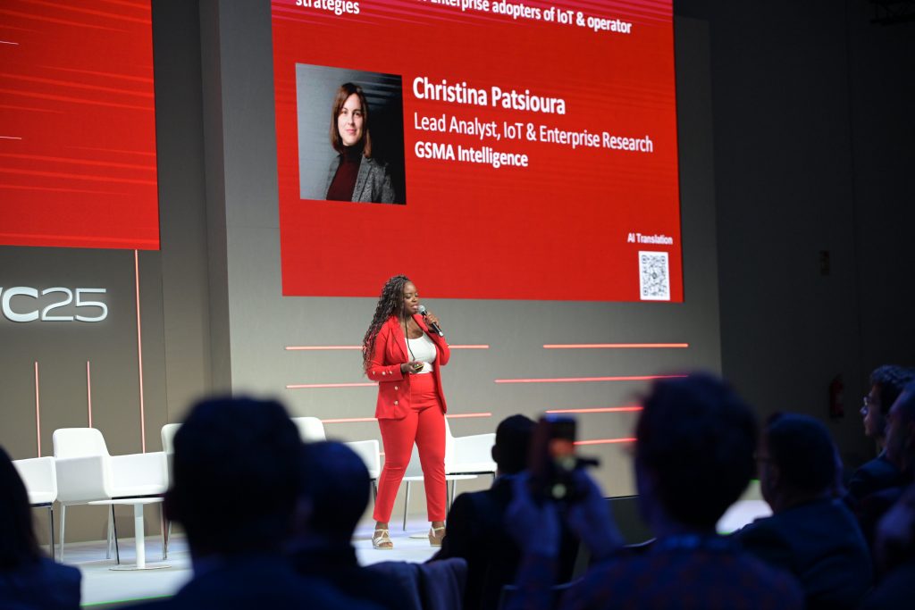 A woman in a red suit speaks on stage holding a microphone. Behind her is a large screen displaying a photo and bio of Christina Patsioura, Lead Analyst at GSMA Intelligence. Audience members are seated in the foreground, facing the stage.