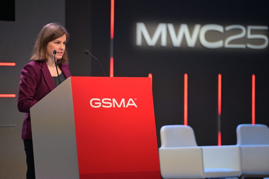 A woman with shoulder-length brown hair, wearing a purple blazer, stands at a podium labeled “GSMA” and speaks into a microphone. Behind her, a dark stage backdrop displays “MWC25” in large white letters and there are a few empty white chairs.