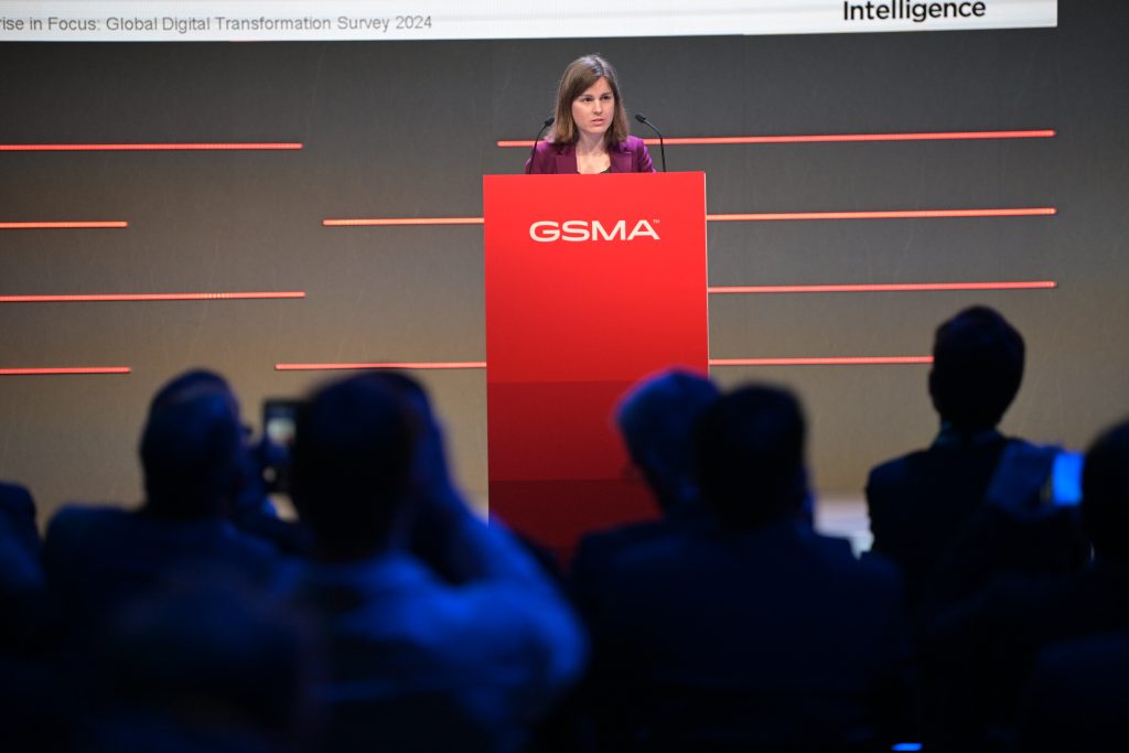 A woman in a purple blazer stands at a red podium with the GSMA logo, addressing an audience. The background features horizontal red lines on a dark wall, and part of a presentation title is visible above her. The audience is seen in shadow.