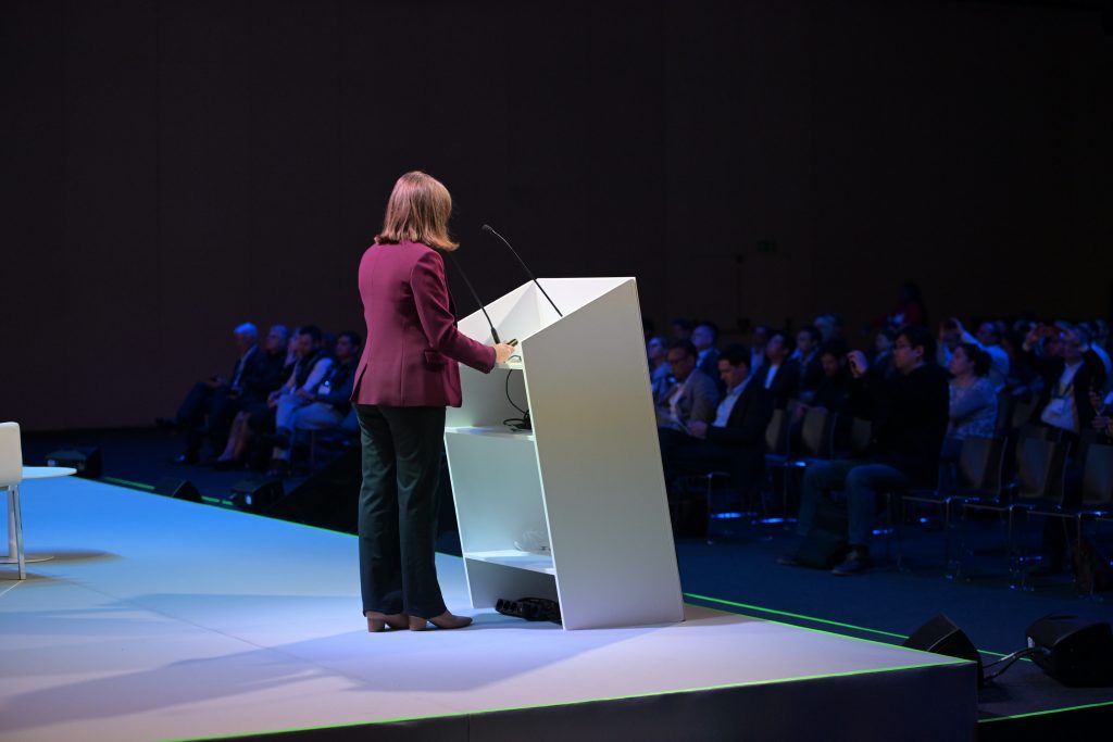 A woman in a maroon blazer and dark pants stands at a white podium on a stage, speaking to an audience seated in rows. The room is dimly lit, with blue accent lighting highlighting the stage area. The focus is on the speaker from behind.