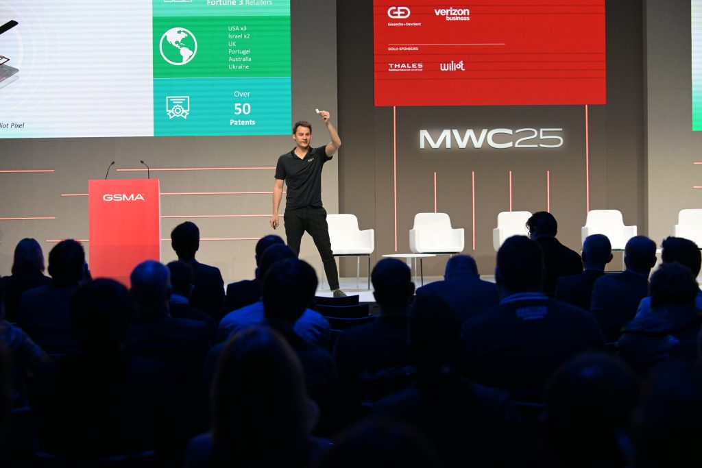 A man in black stands onstage, gesturing with one hand, addressing a seated audience at a technology event. Behind him are a red GSMA podium, white chairs, and large screens displaying charts, company logos, and “MWC25.”.