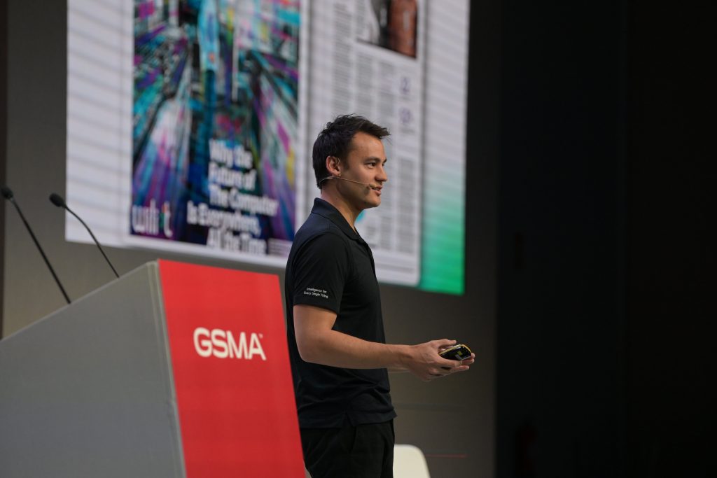 A man wearing a black shirt and headset microphone is speaking on stage beside a podium with “GSMA” in white letters. Behind him, a large screen displays colorful graphics and text, suggesting a tech conference or presentation setting.