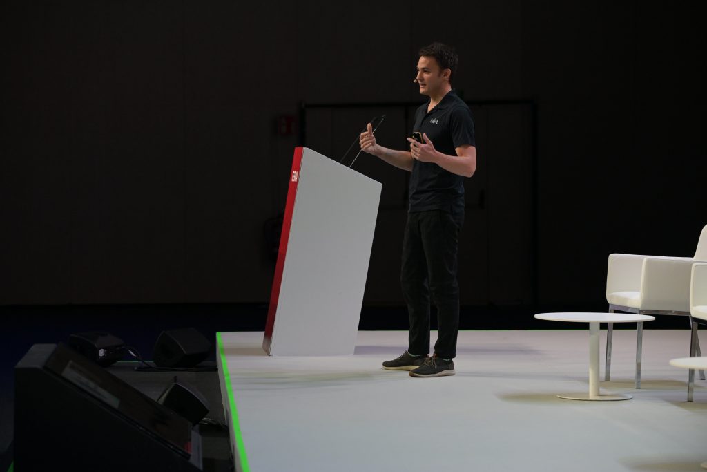 A person in a black shirt and pants stands on stage behind a white podium, speaking and gesturing with one hand. The background is dark, with empty white chairs and a spotlit stage area to the right. The atmosphere suggests a formal presentation or talk.