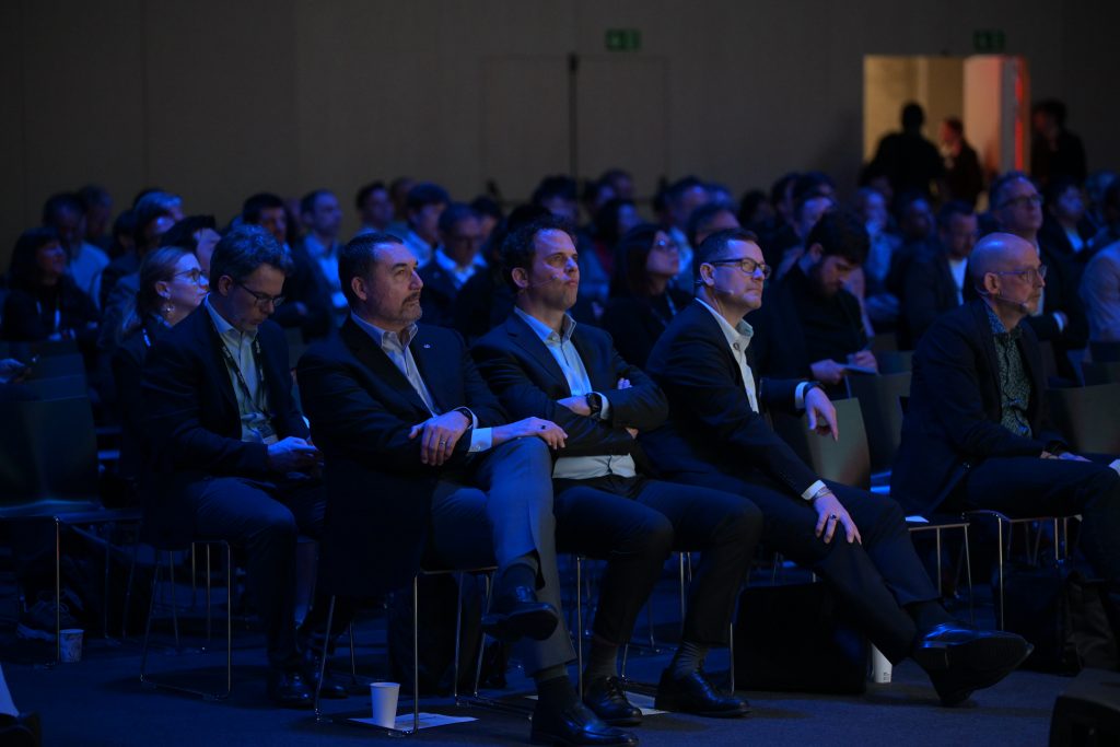 Audience of mostly men in suits sit attentively in rows of chairs at a dimly lit conference or seminar; some have arms crossed and faces focused, with others in the background also listening. Blue lighting highlights the front row.