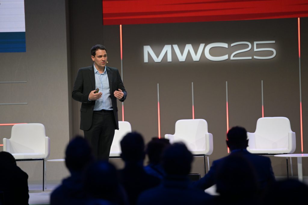 A man in a black suit gives a presentation on stage at MWC25. He stands in front of four empty white chairs, holding a remote, with “MWC25” displayed on a large screen behind him. Several silhouetted audience members watch him.