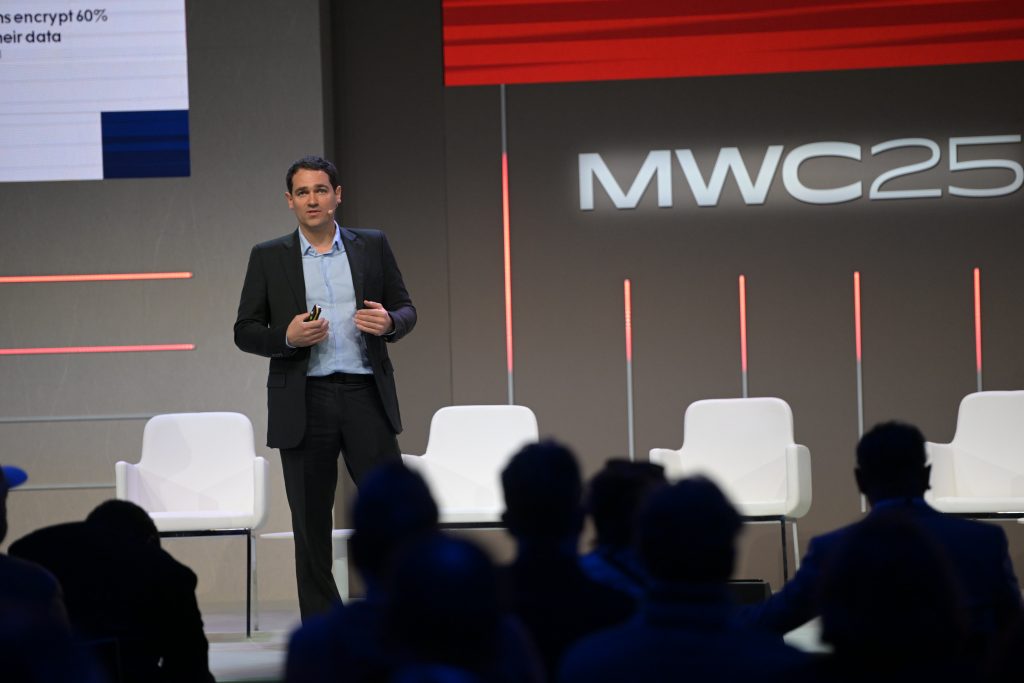 A man in a suit stands on a stage giving a presentation at MWC23. He holds a small device, possibly a remote or microphone. Four empty white chairs are behind him, and an audience is visible in silhouette. The background features the MWC23 logo and vertical light bars.