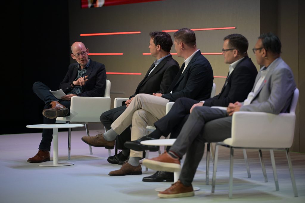 Five men in business attire sit on modern white chairs on a stage. One man on the left, holding papers, faces and speaks to the other four, who are sitting in a row facing him. Red horizontal lines decorate the background wall.