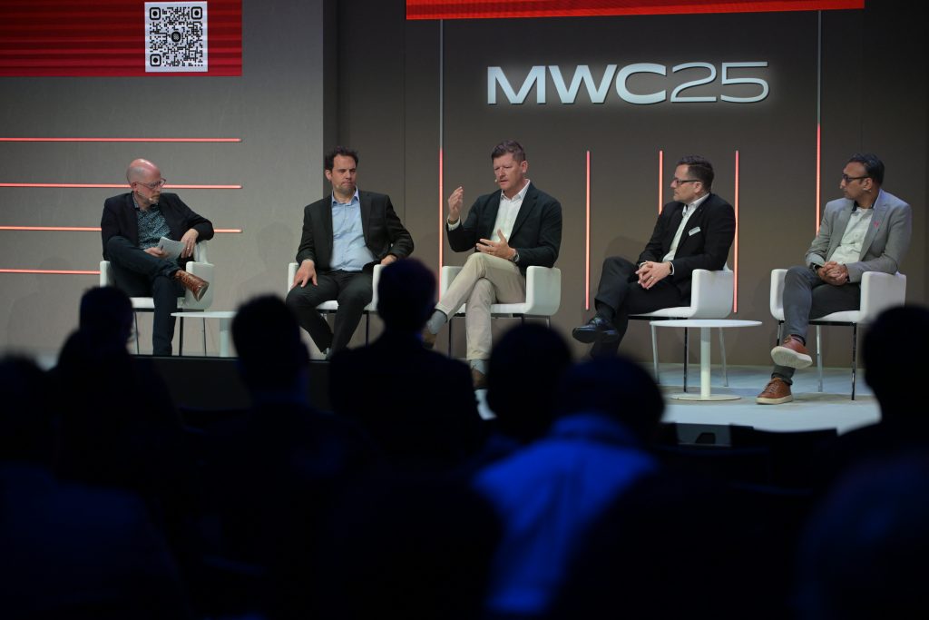 Five men in business attire sit on a stage panel at MWC25, engaged in discussion. The man in the center gestures while speaking. A large "MWC25" sign is on the wall behind them. Audience silhouettes are visible in the foreground. A QR code is seen on the upper left.