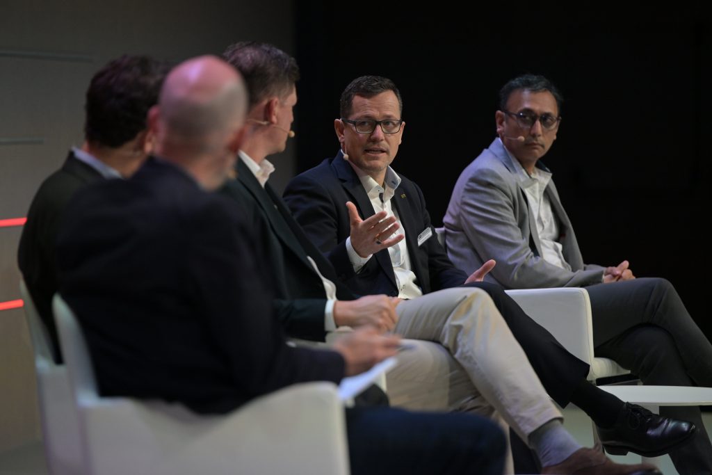 Five men sit on a panel stage, dressed in business attire. The central man, gesturing with his hand, appears to be speaking, while the others listen attentively. The background and seating are minimalistic, emphasizing the discussion.