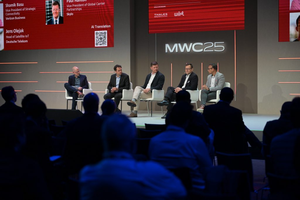 A panel of five men in business attire sit onstage discussing at MWC25, with a red digital event screen behind them. Audience members watch from the foreground, seated in a dimly lit conference hall.