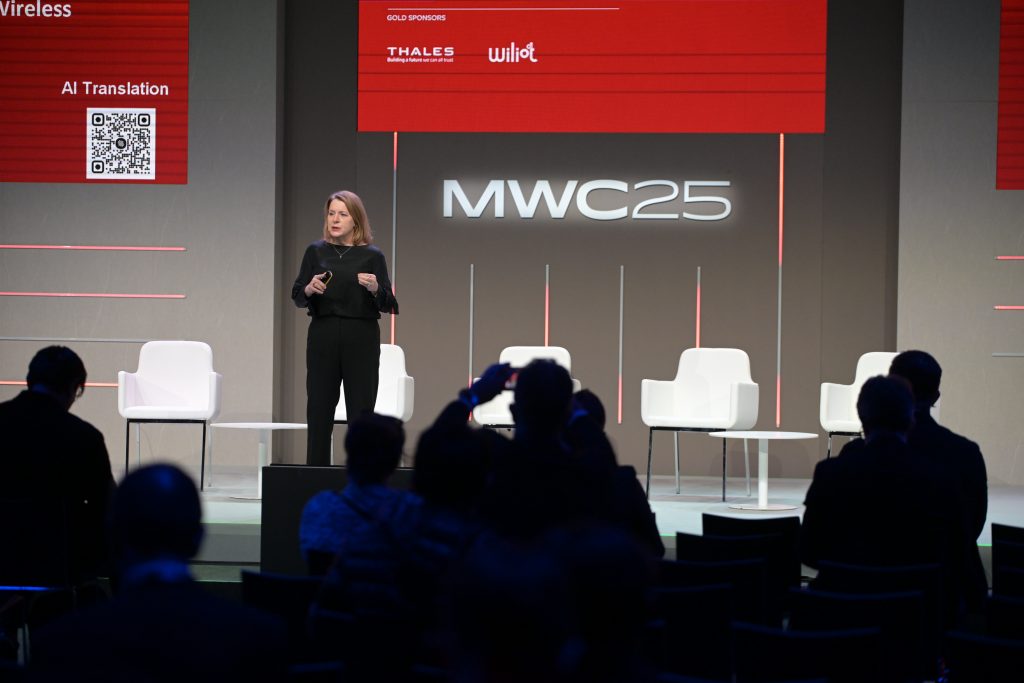 A woman in black speaks on stage at MWC25 in front of empty white chairs. Audience members sit and take photos. Red banners overhead display sponsor logos and a QR code. The background is gray with the “MWC25” logo illuminated at center.