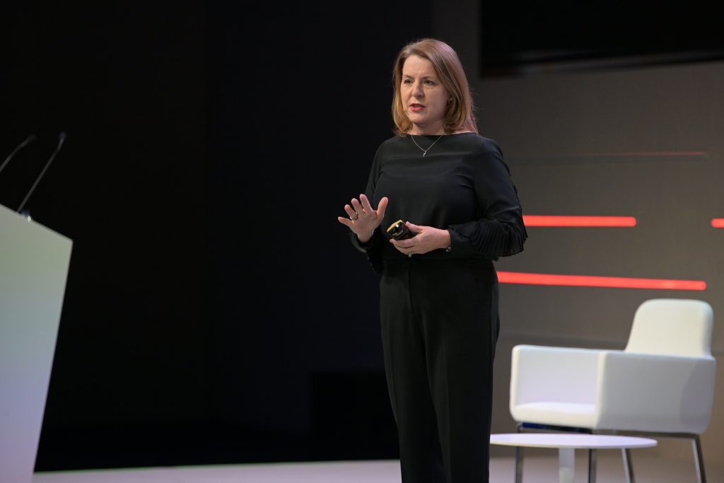 A woman with shoulder-length light brown hair, wearing a black long-sleeve outfit, stands on stage speaking. She gestures with one hand, holding a small object in the other. A white chair and podium are on stage, with red horizontal lines in the background.